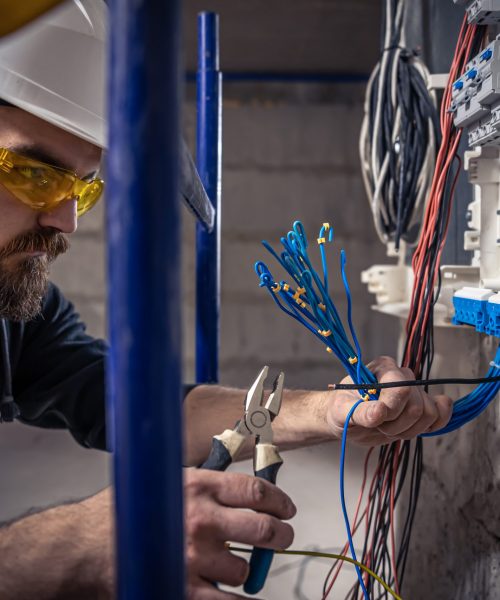 A male electrician works in a switchboard with an electrical connecting cable, connects the equipment with tools.